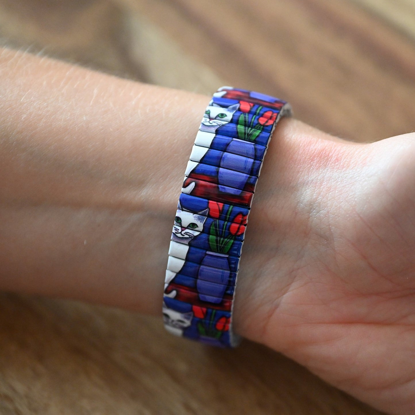 Farbenfroher Armreif mit Katzenmotiv und Blumen, getragen am Handgelenk. Blaue, weiße und rote Details auf flexiblem Band.