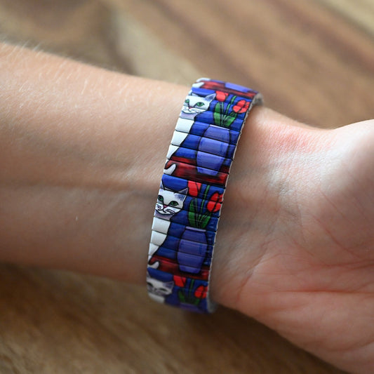 Farbenfroher Armreif mit Katzenmotiv und Blumen, getragen am Handgelenk. Blaue, weiße und rote Details auf flexiblem Band.