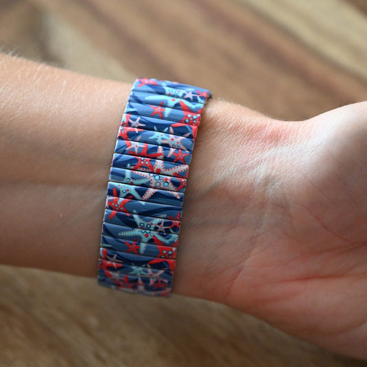 Blaues Stretcharmband mit buntem Seestern-Muster am Handgelenk, auf Holzuntergrund fotografiert