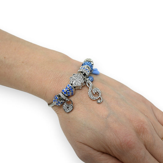 Rigides Charm-Armband mit blauen und silbernen Anhängern, darunter ein Notenschlüssel, Herz und Kleeblatt auf einem Handge...