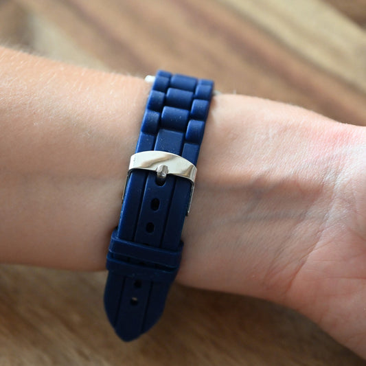 Blaues Silikonarmband mit silberner Schnalle am Handgelenk, auf Holzuntergrund. Nahaufnahme des Verschlusses.