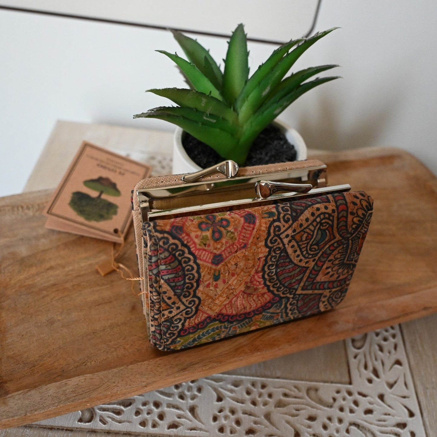 Kompakte Brieftasche aus Kork mit buntem Paisleymuster auf Holztisch neben Sukkulente und Buch, Bohemian-Stil