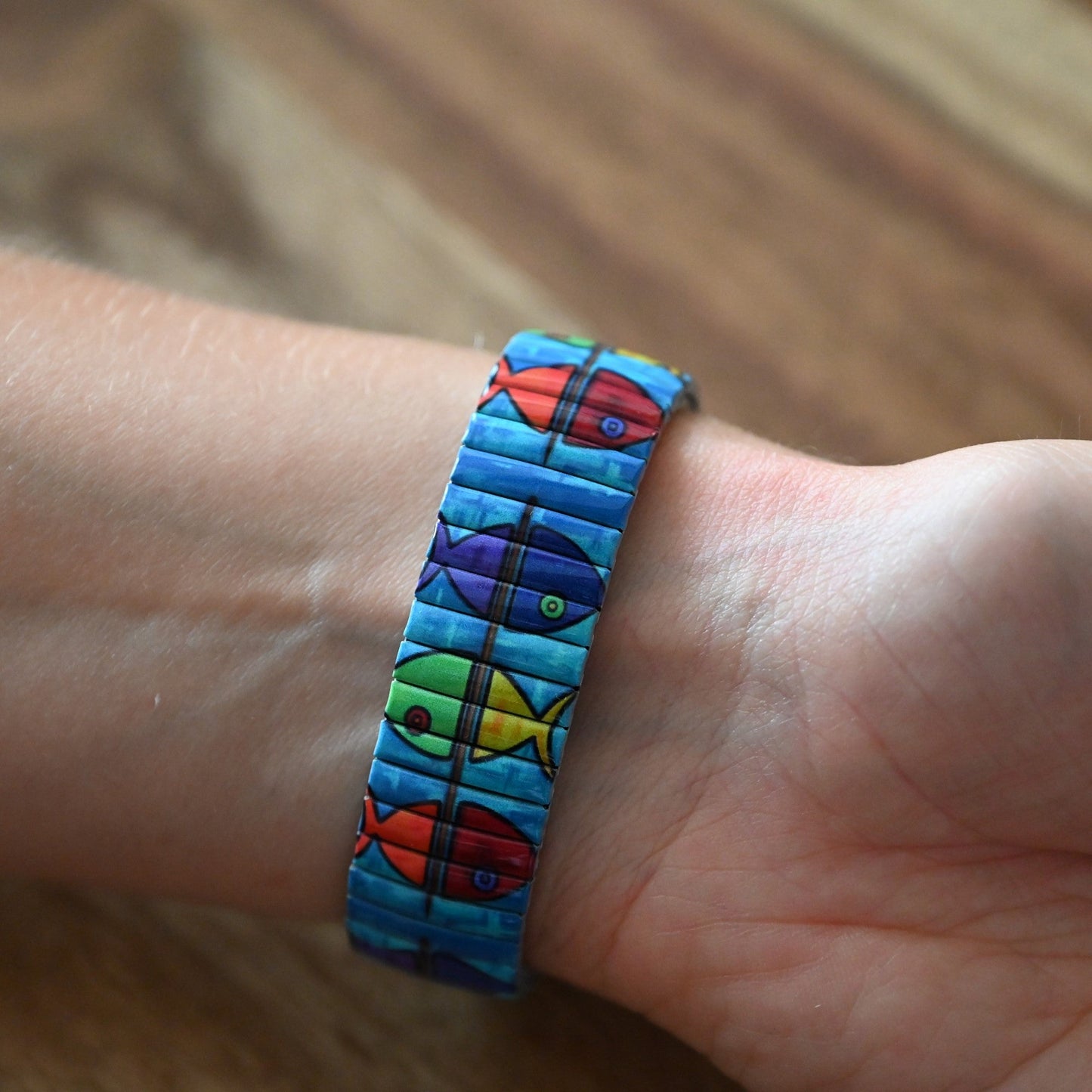 Buntes Armband mit Fischmotiven in Blau, Rot, Lila und Grün auf einem Handgelenk vor Holzhintergrund