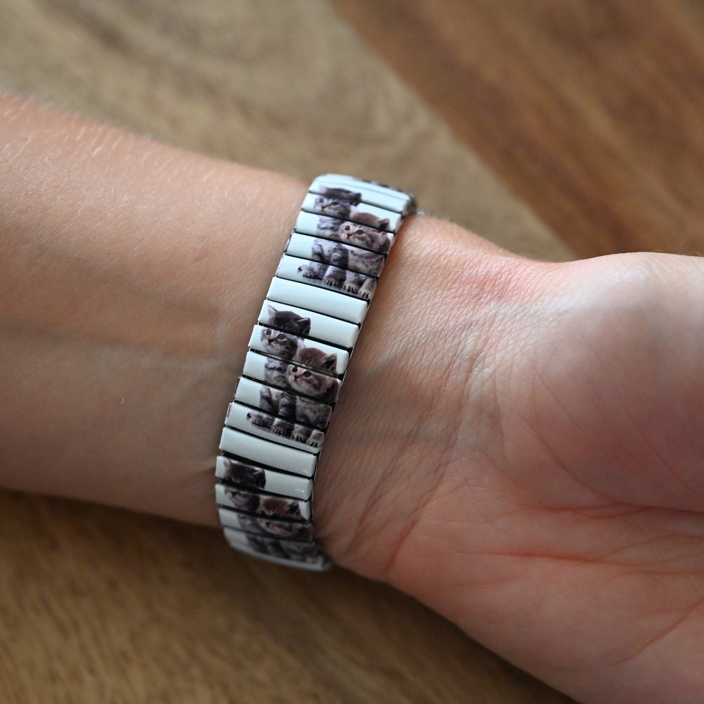 Erweiterbare Armbanduhr mit Katzenbildern auf dem Band, getragen an einem schlanken Handgelenk auf Holzuntergrund