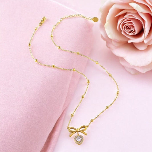 Zarte goldene Halskette mit Schleifenanhänger und Herz auf rosa Hintergrund neben einer Rose, elegantes Schmuckstück