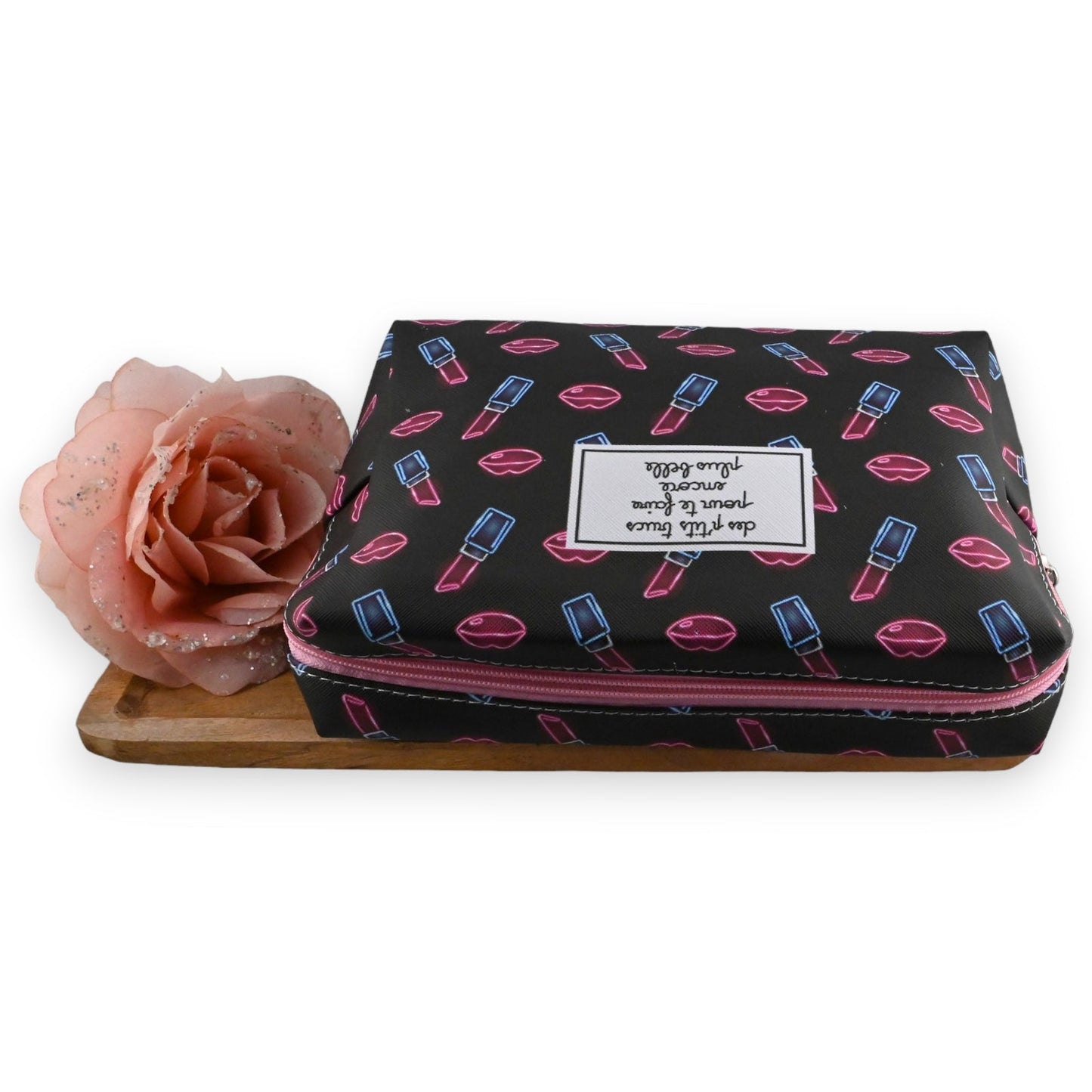 Schwarze Kosmetiktasche mit Lippenstift- und Lippen-Motiv, rosa Reißverschluss, daneben eine rosa Rose auf Holzunterlage