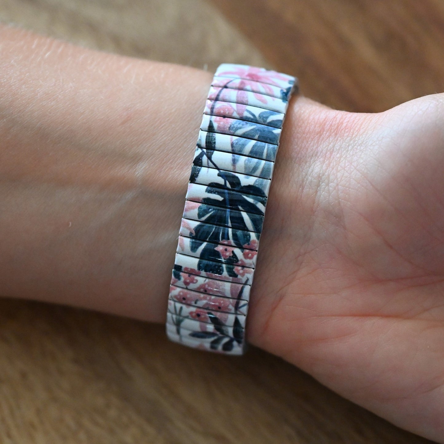 Elastisches Armband mit tropischem Blumenmuster in Weiß, Rosa und Blau, getragen am Handgelenk auf Holzuntergrund