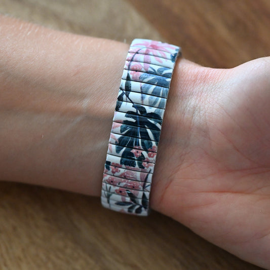Elastisches Armband mit tropischem Blumenmuster in Weiß, Rosa und Blau, getragen am Handgelenk auf Holzuntergrund