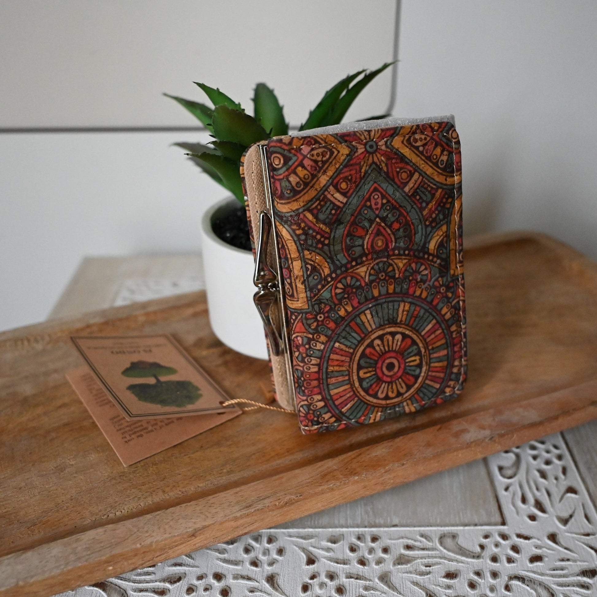 Bunte kompakte Kork-Geldbörse mit orientalischem Muster neben Pflanze und Holztag auf rustikalem Holzbrett