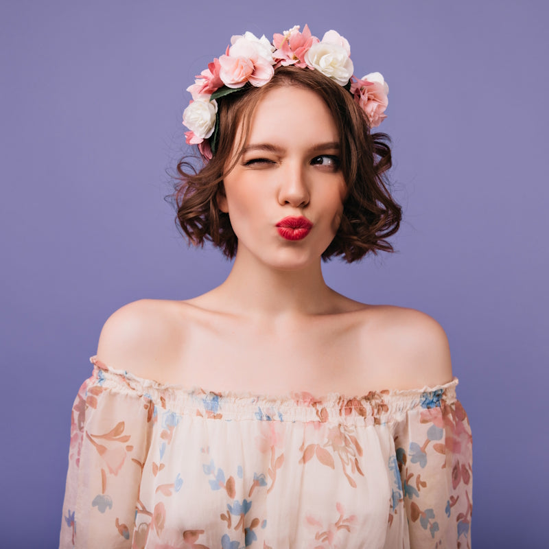 Frau mit Blumenkranz aus rosa und weißen Rosen, schulterfreies Blumenkleid, Kussmund vor violettem Hintergrund