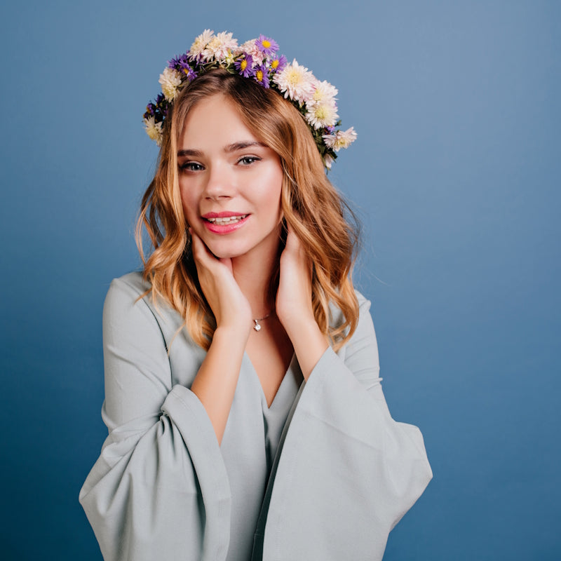 Lächelnde blonde Frau mit buntem Blumenkranz, Hände am Hals, graues Sakko vor blauem Hintergrund