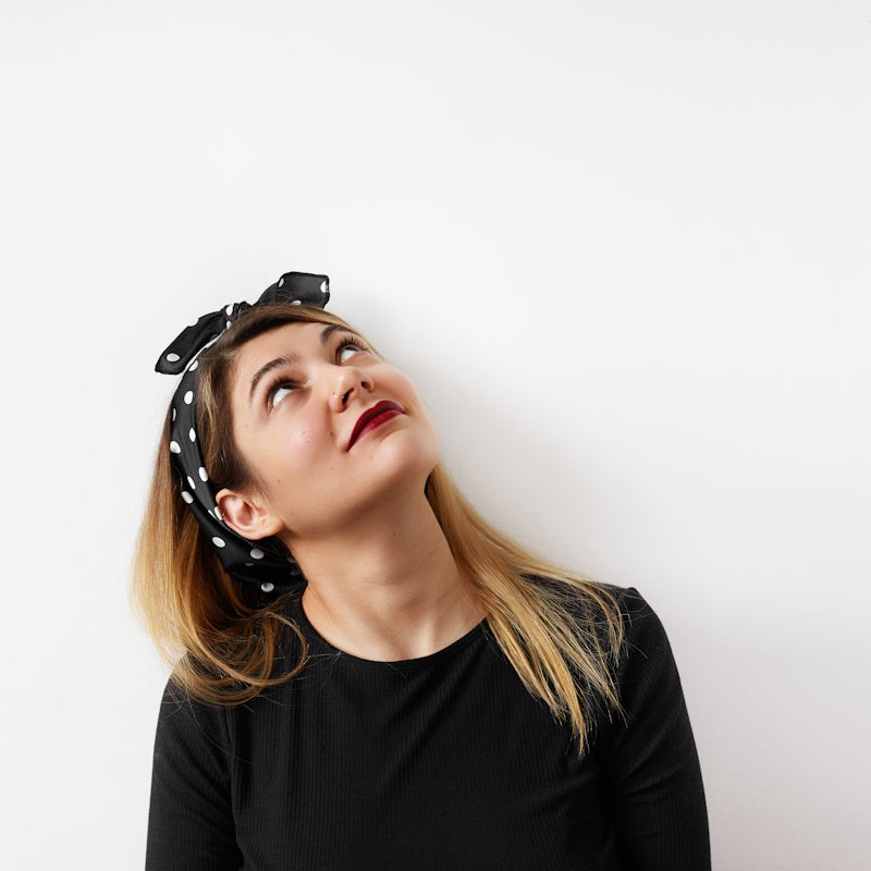 Junge Frau mit schwarzem Polka-Dot-Haarband und schwarzem Pullover, blickt nachdenklich nach oben vor weißem Hintergrund