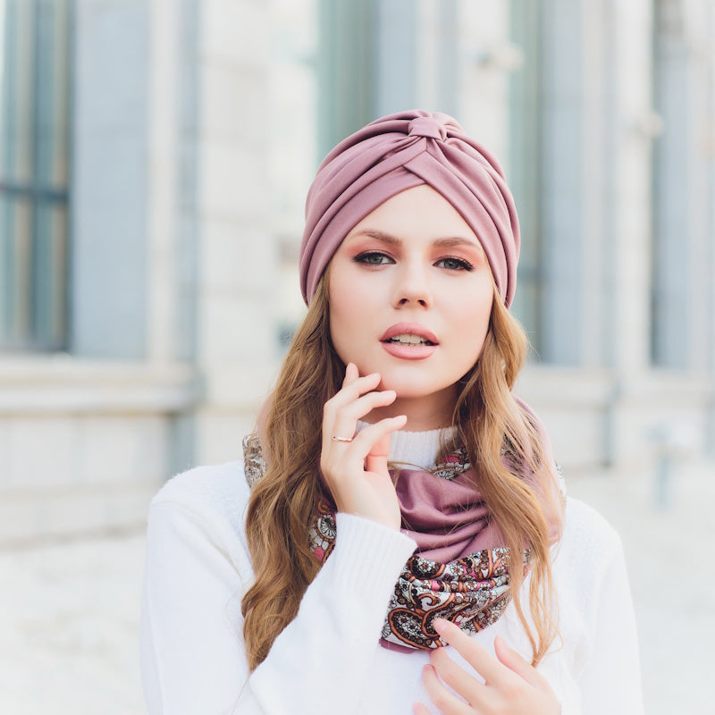 Elegante Frau mit rosa Turban und gemustertem Schal, Hand am Gesicht, vor klassischer Säulenarchitektur