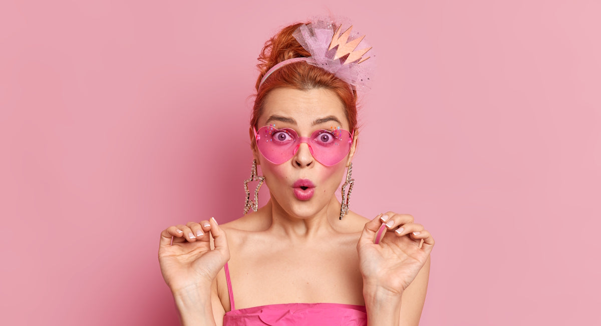 Überraschte Frau mit herzförmiger rosa Sonnenbrille und goldener Krone, offener Mund vor rosa Hintergrund