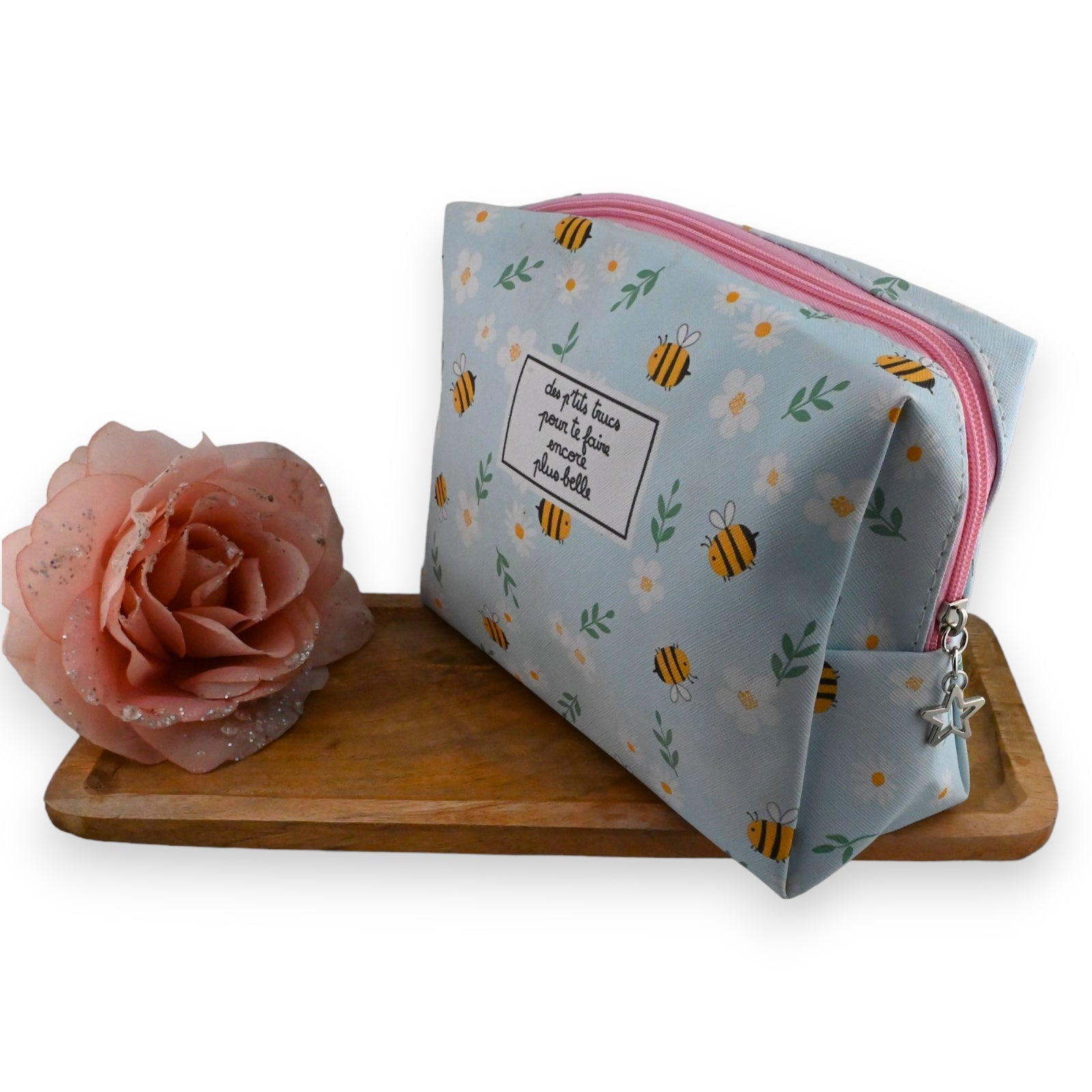 Toilettentasche mit Bienenmuster, rosa Reißverschluss und Sternanhänger neben rosa Rose auf Holzbrett